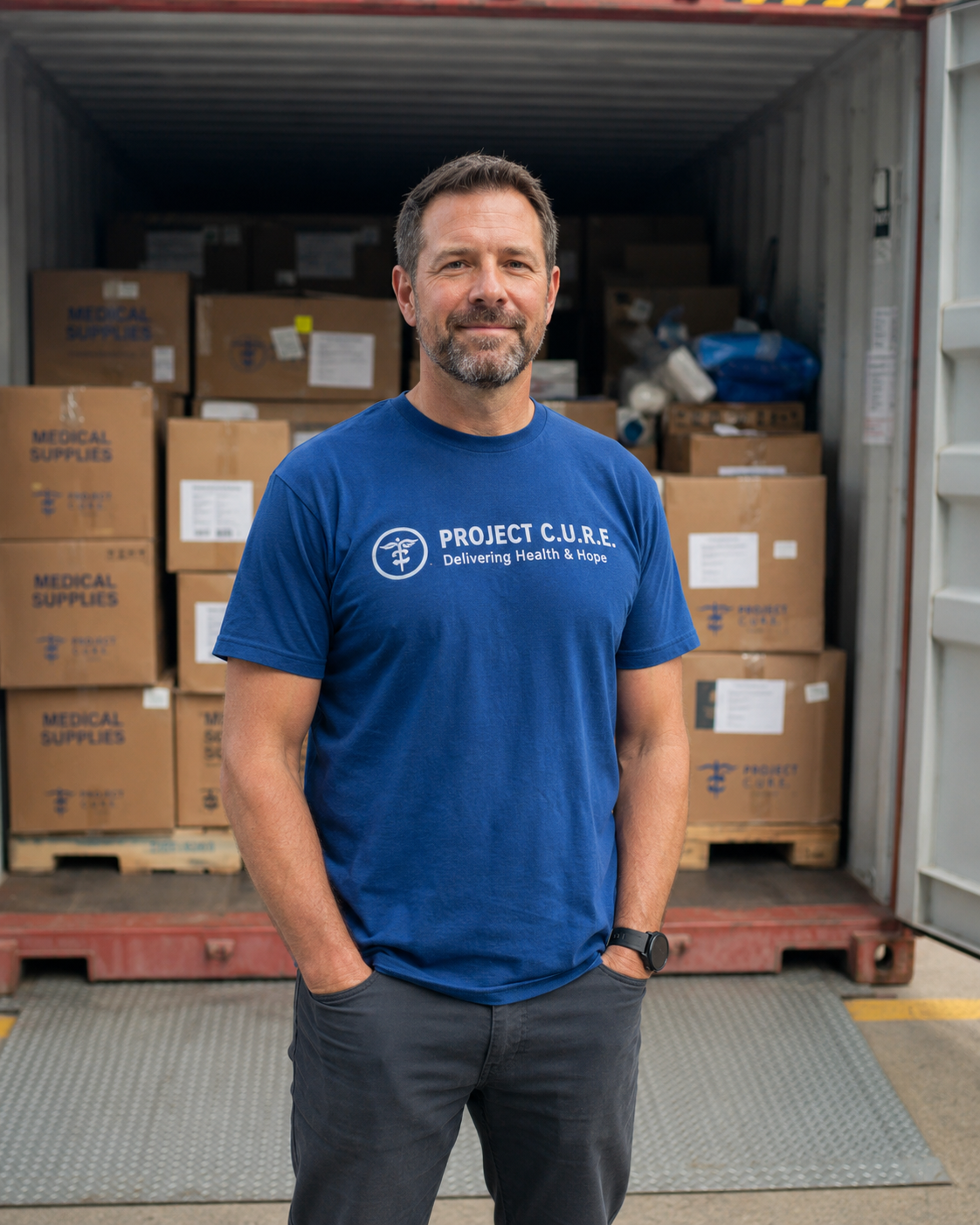 A Project C.U.R.E. volunteer standing before a shipping container loaded with boxes of donated medical supplies