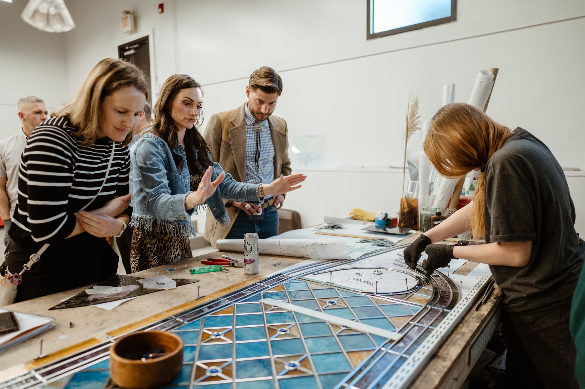Scottish Stained Glass team and visitors gathered around a craftsperson assembling a stained glass panel in the workshop.